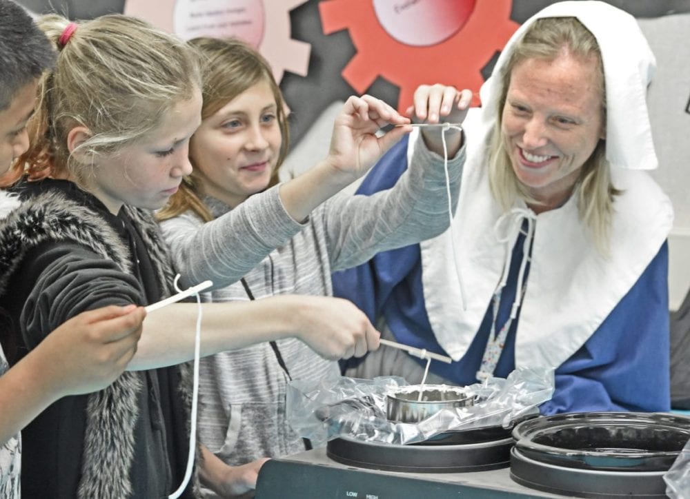 Fifth-grade teacher, Melinda Katambwa, right,  dressed as a Colonial era woman, assists fifth-graders Kiley Norman, left, and Lily Drummond as they dip string into hot wax to make a candle while participating in Colonial Day at  Newhall elementary School in Newhall on Friday.  Dan Watson/The Signal