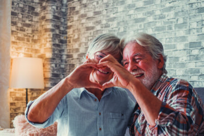 Close up of hands of two mature grandparents making a heart shape with their fingers together. Smiling and having fun in love people.