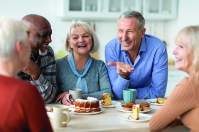 Multiracial group of happy senior people sitting around table drinking tea with cake and having conversation, smiling and laughing, having home party or enjoying time at nursing home