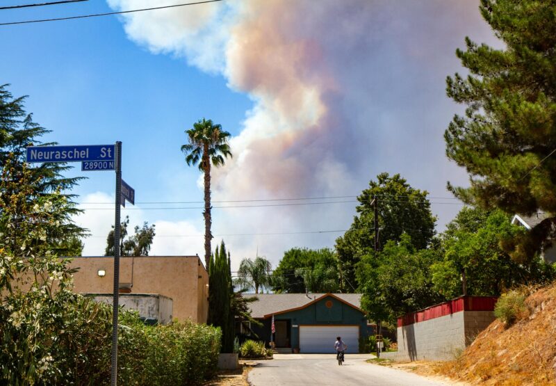 A boy rides his bike in Val Verde, an unincorporated area of the Santa Clarita Valley, with a cloud of smoke from the Canyon Fire lingering overhead on Aug. 7, 2025. Katherine Quezada/The Signal