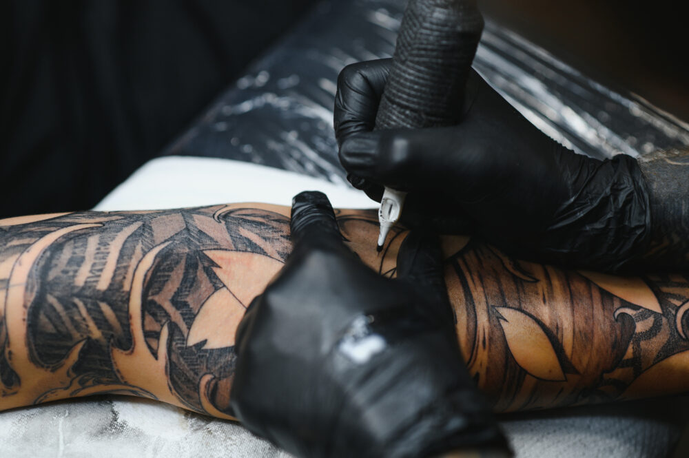 Cropped close up of a bearded tattoo artist working at his studio tattooing sleeve on the arm of his male client. Man getting tattooed by professional tattooist. Adobe Stock photo.