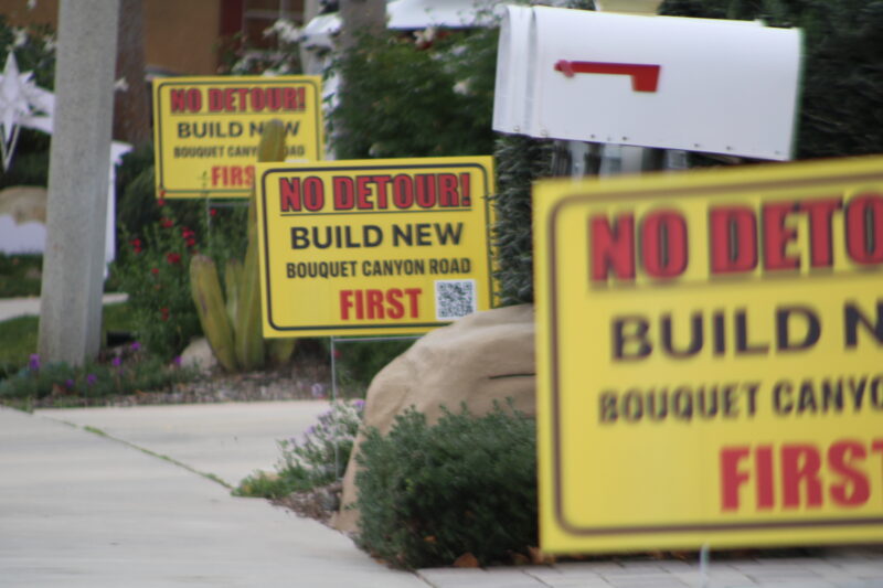 Signs on three consecutive lawns along Woodside Drive on Tuesday, Dec. 23, 2025, calling for the cancellation of a planned detour that will route drivers away from Bouquet Canyon Road for 11 months.