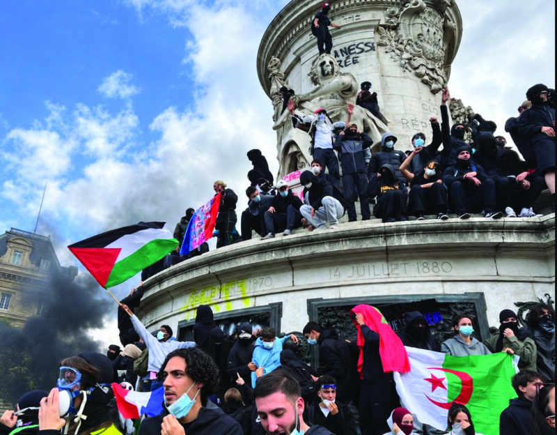 Masked protesters gather at the Monument to the Republic at Place de la République in Paris during far-left demonstrations on Oct. 17, 2026, with Moroccan, Algerian, and Palestinian flags visible as thick black smoke rises.