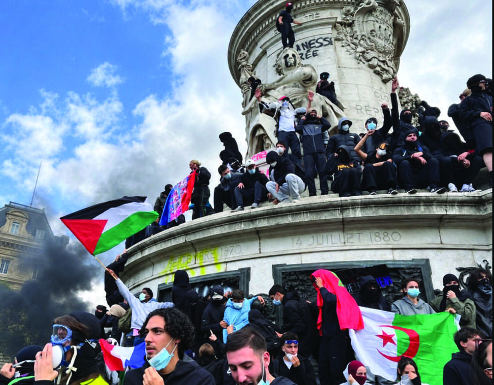 Masked protesters gather at the Monument to the Republic at Place de la République in Paris during far-left demonstrations on Oct. 17, 2026, with Moroccan, Algerian, and Palestinian flags visible as thick black smoke rises.